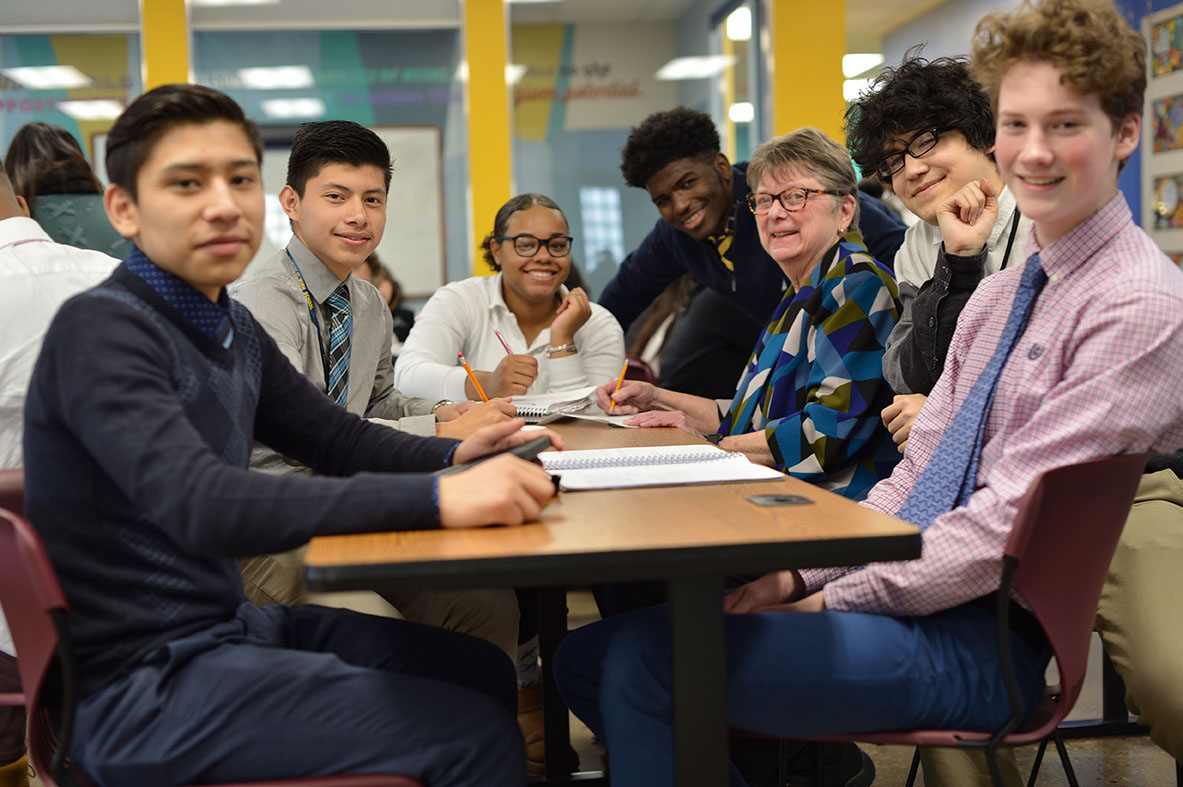 A group of diverse students and one adult sit around table and smile towards camera